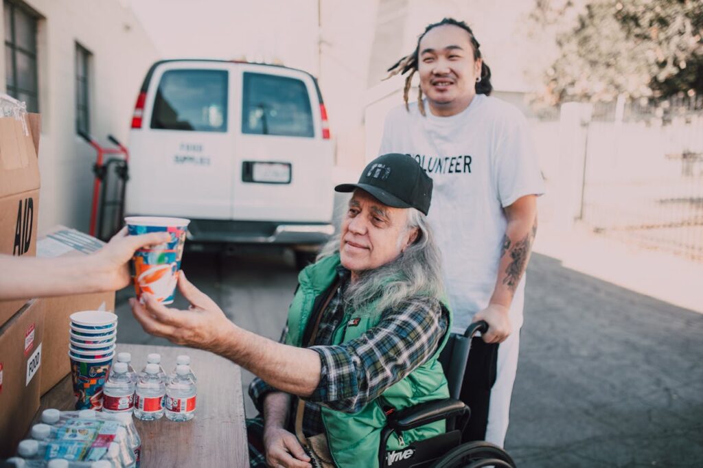 A volunteer assists an elderly man in a wheelchair receiving aid outdoors.